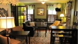 A cozy living room with a sofa, armchairs, a coffee table, and a dining table. There is a large window with curtains, a fireplace, and a lamp. - Monadnock Inn (Jaffrey)