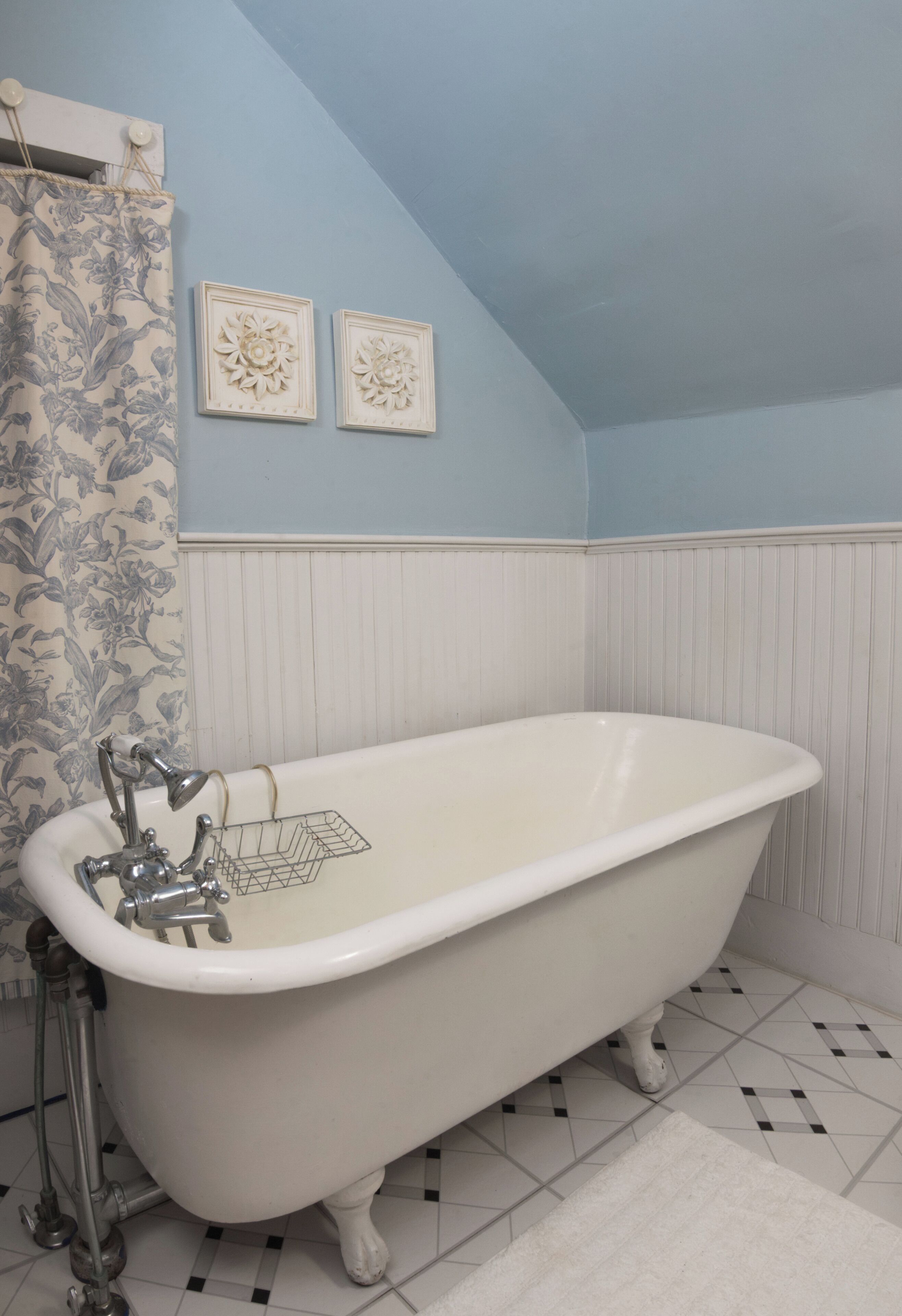 Un baño con bañera tipo patas de león, suelo de baldosas blancas, papel tapiz floral y dos adornos florales en la pared.