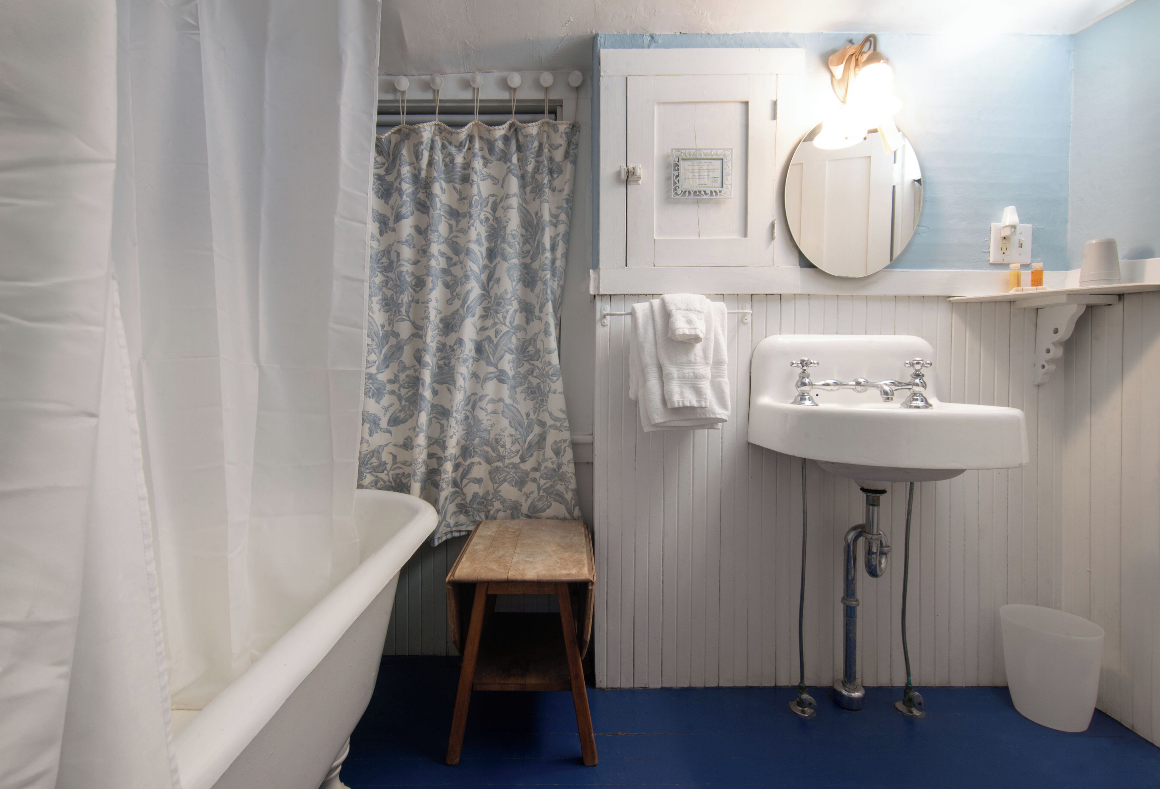 A bathroom with a bathtub, a sink, a shower curtain, a small wooden stool, and a white trash bin.