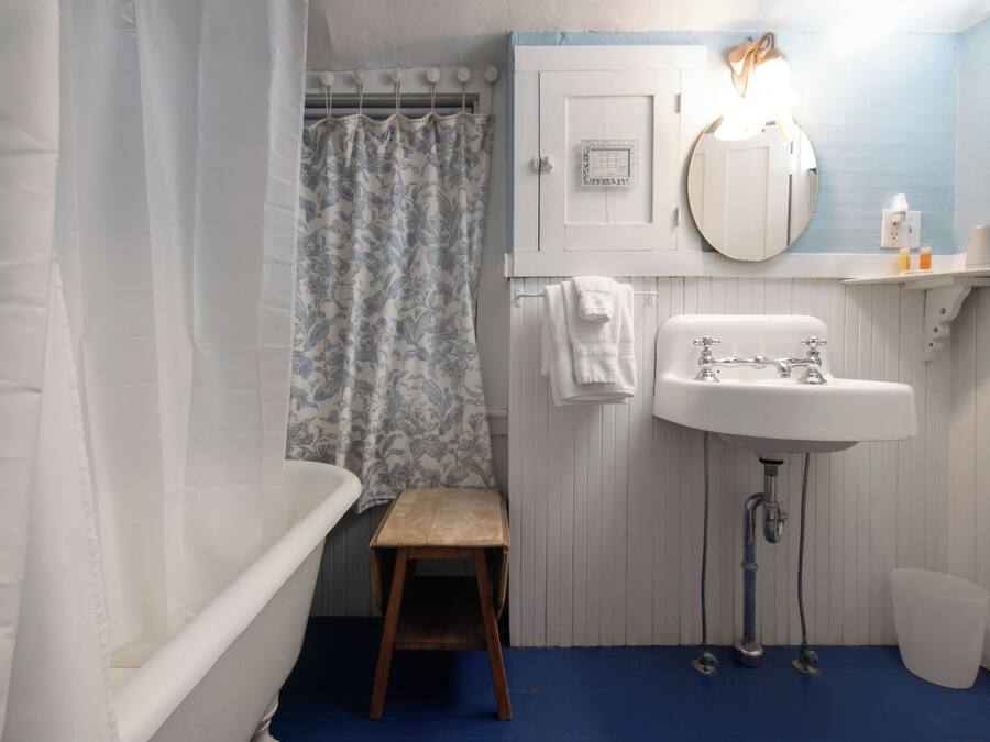 A bathroom with a bathtub, a sink, a shower curtain, a small wooden stool, and a white trash bin.