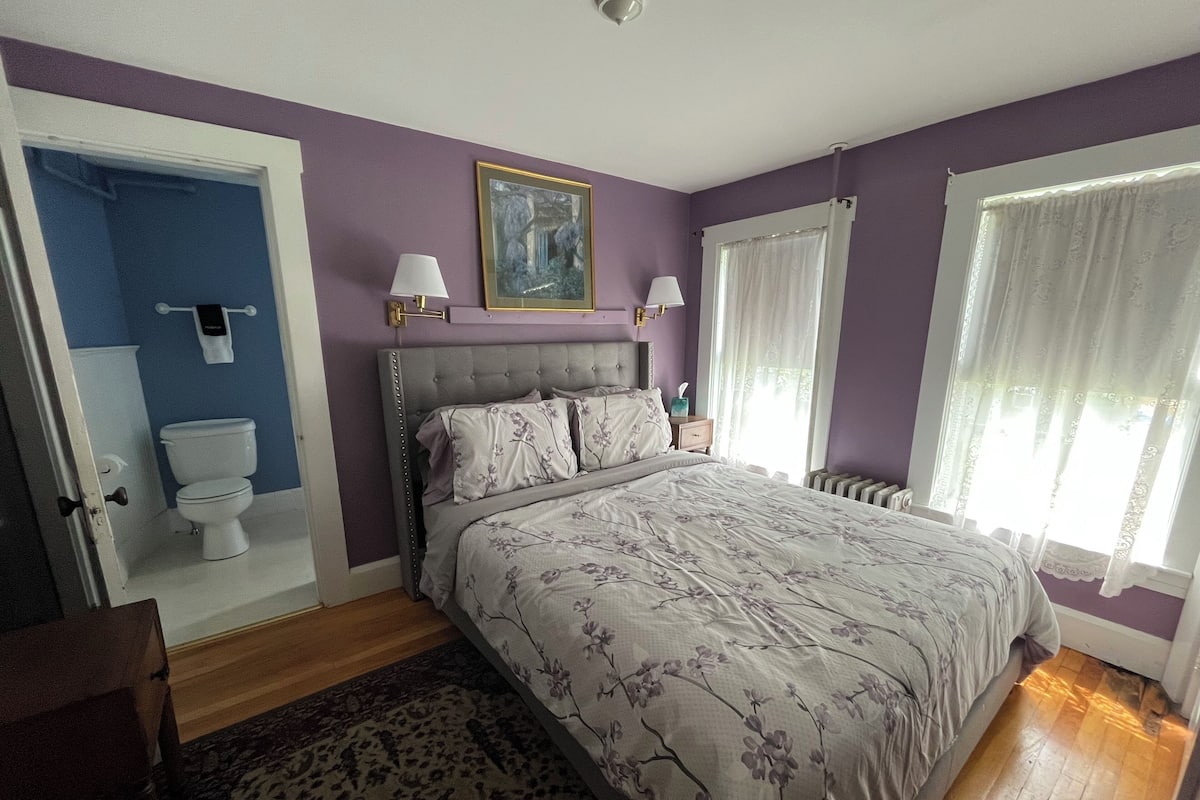 A bedroom with a bed, a bedside table, a nightstand, a window with curtains, a radiator, a door leading to a bathroom, and a wall-mounted painting.
