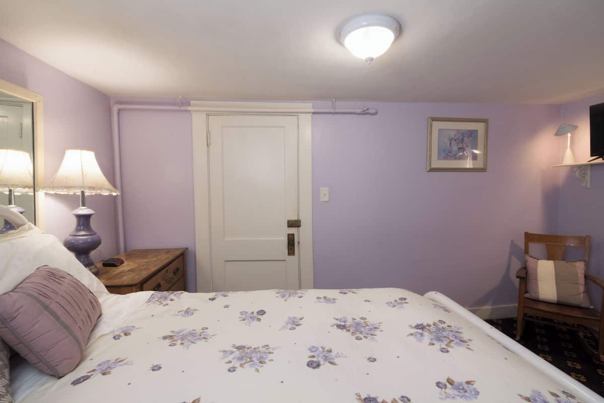 A bedroom with a bed, a nightstand, a chair, a door, a wall-mounted TV, and a ceiling light.