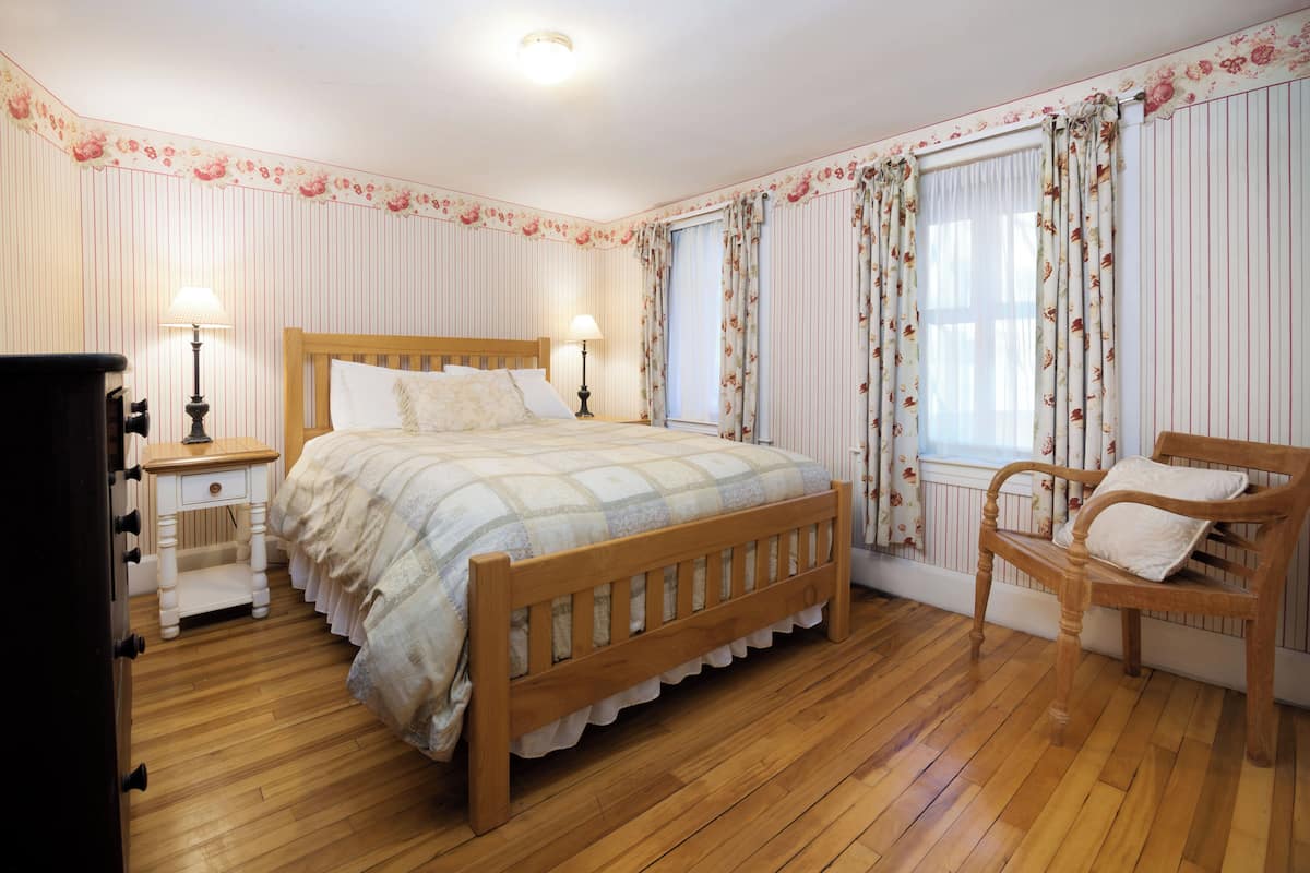 A bedroom with a wooden bed, two bedside tables, a chair, and a window with floral curtains.