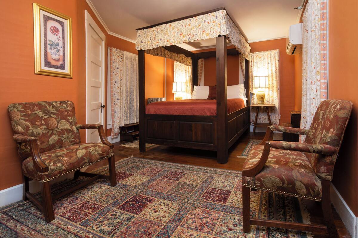 A traditional bedroom with a four-poster bed, two upholstered chairs, a patterned rug, and floral curtains.