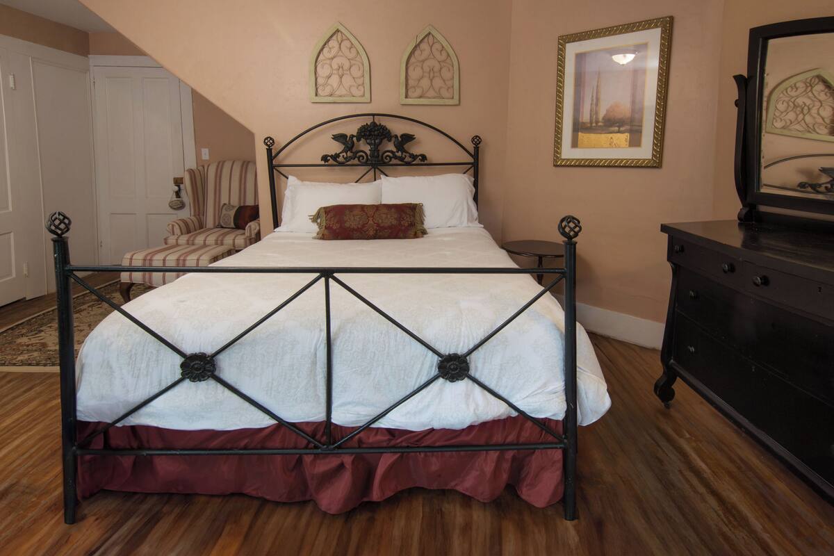 A bedroom with a four-poster bed, a wooden floor, a dresser, and a chair.