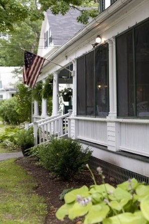 A white house with a porch, an American flag, and a garden with green plants.