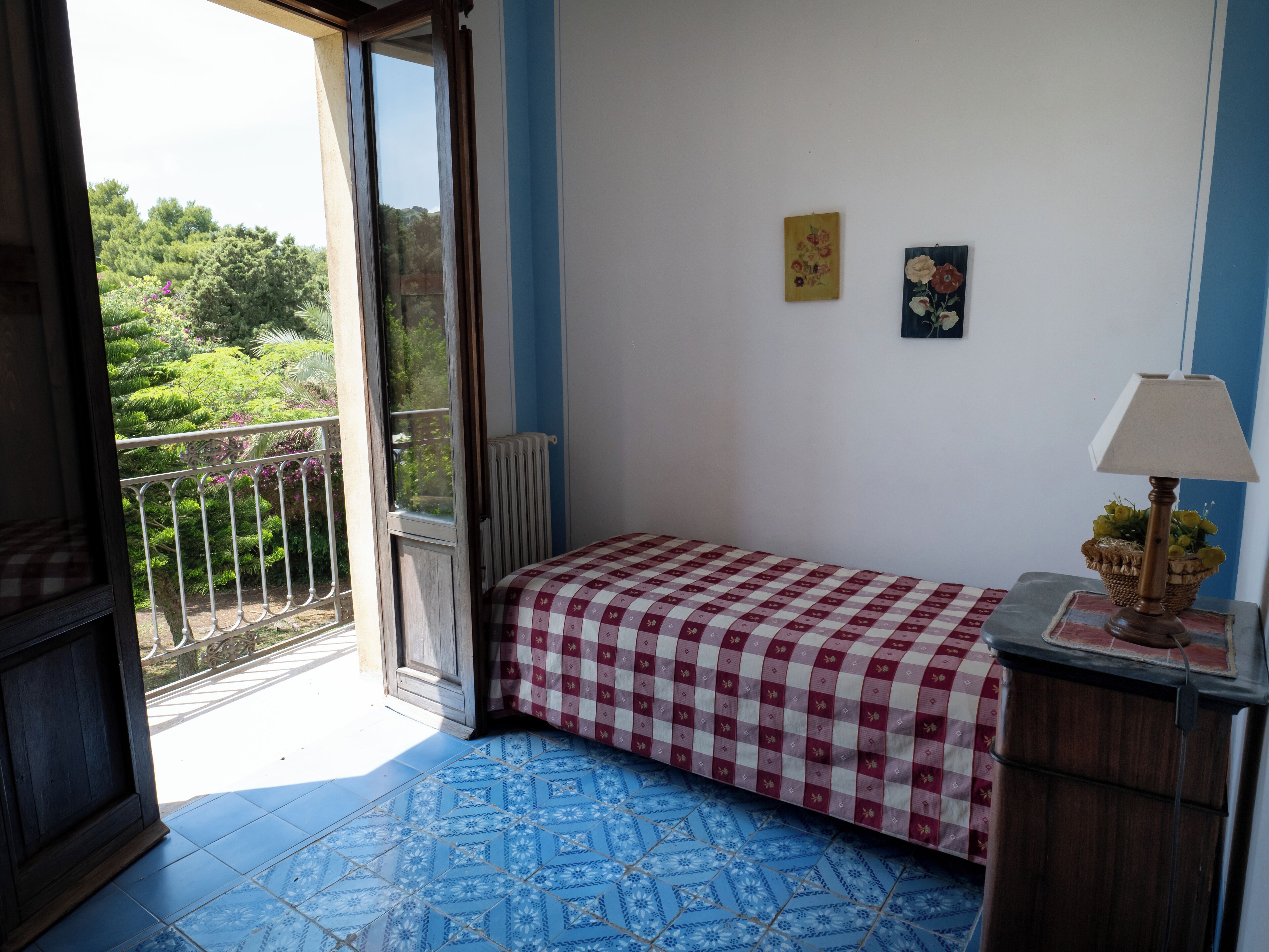 Camera da letto con copriletto a quadri, un termosifone, un tavolino con una lampada e vista sul verde attraverso una finestra.