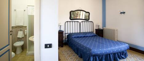 A bedroom with a bed, a bedside table, a lamp, a radiator, and a bathroom visible through an open door.