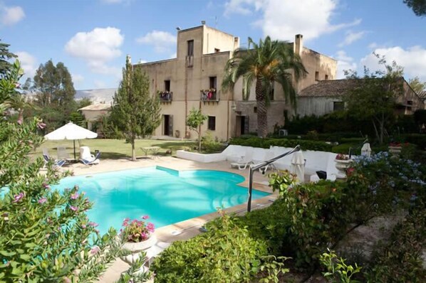 A pool area with a clear blue pool, surrounded by lush greenery and a building with balconies. - Agriturismo Baglio Fontana (Buseto Palizzolo)
