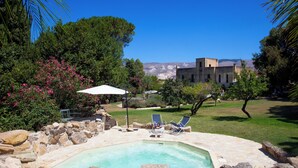 A swimming pool surrounded by rocks, with two lounge chairs and a white umbrella. - Agriturismo Baglio Fontana (Buseto Palizzolo)