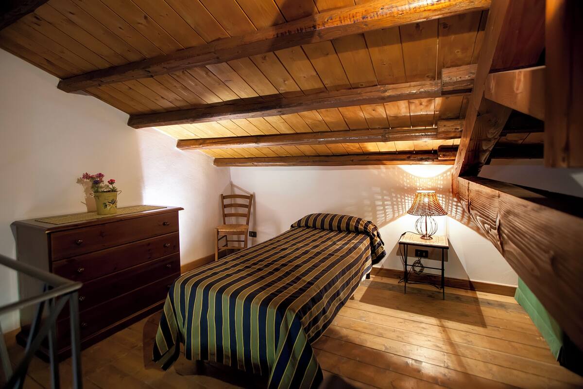Een slaapkamer met een houten plafond, een bed met gestreept beddengoed, een dressoir, een stoel en een tafel met een lamp.
