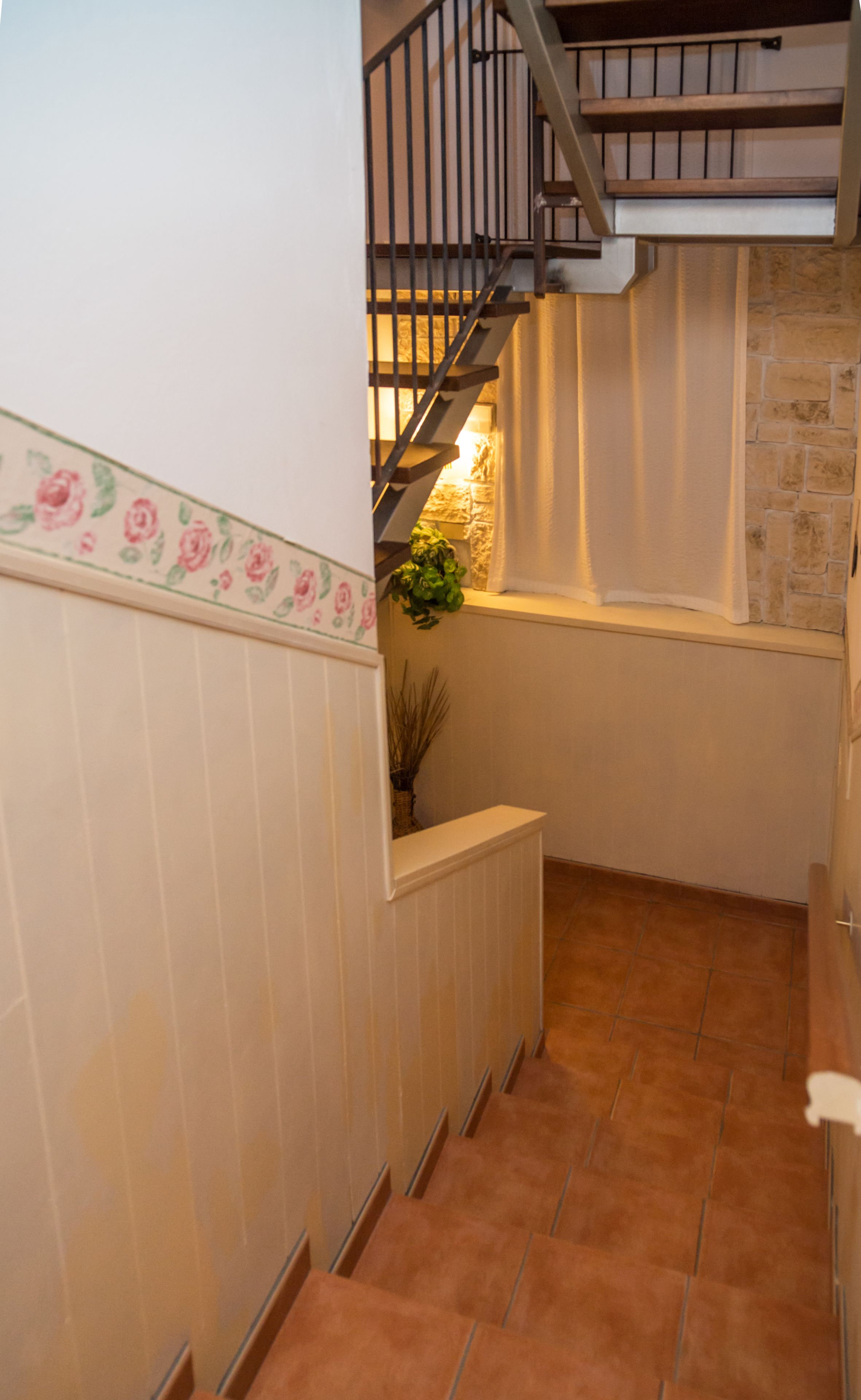 Un couloir étroit doté d’un sol carrelé, d’un escalier avec une rampe en métal et d’un mur orné d’un motif floral.