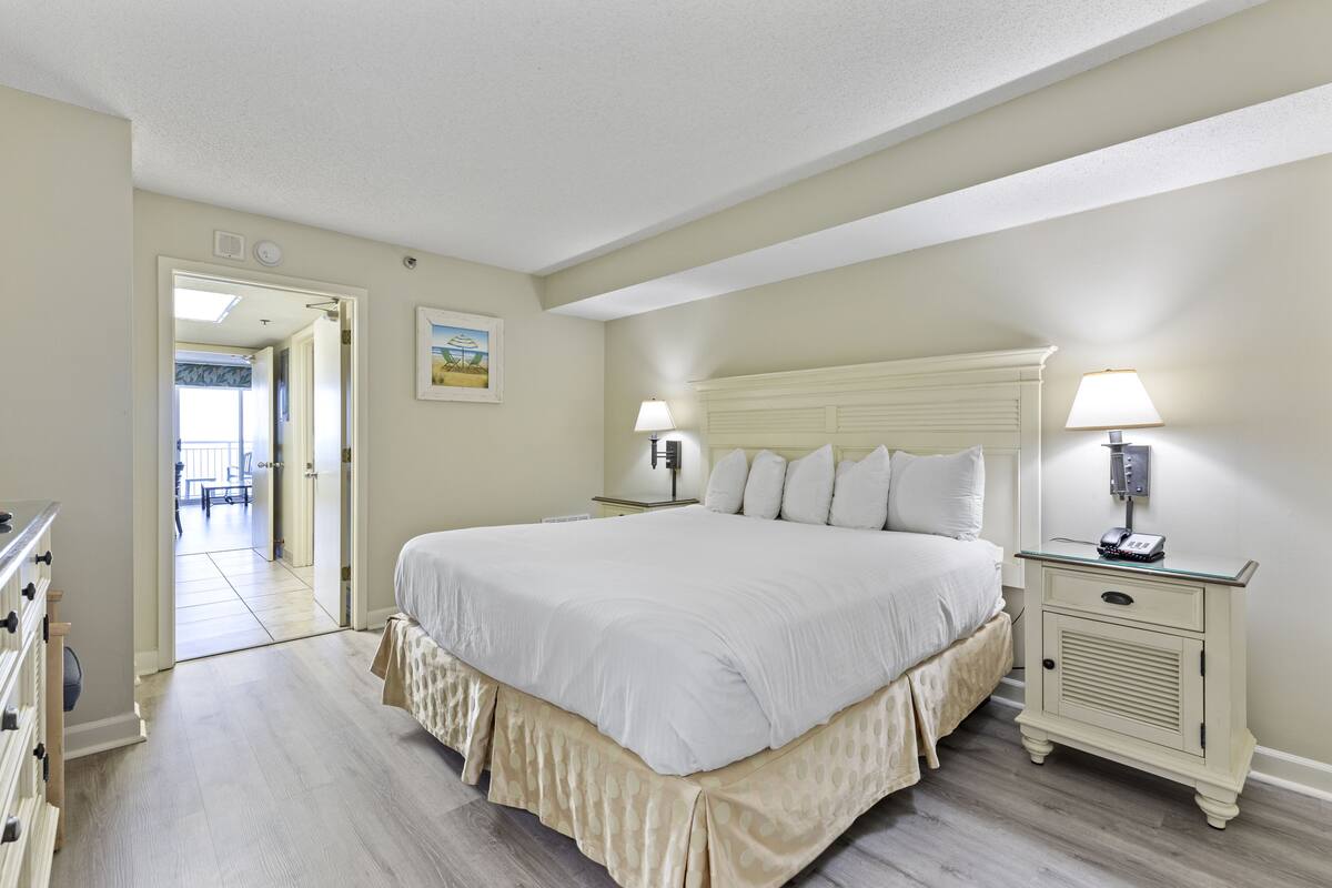 Un dormitorio con una cama grande, mesitas de noche, un aparador y una puerta que da a otra habitación.
