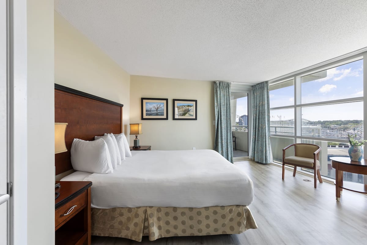 Habitación de hotel con una cama grande, una mesita de noche de madera, dos cuadros enmarcados en la pared, un ventanal con vistas a la ciudad y un balcón con una silla.