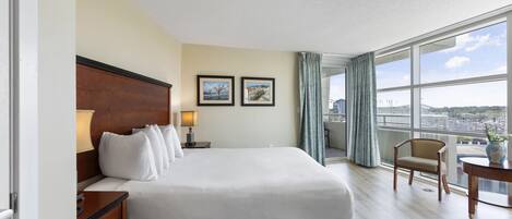 A hotel room with a large bed, a wooden nightstand, two framed pictures on the wall, a window with a view of the city, and a balcony with a chair.
