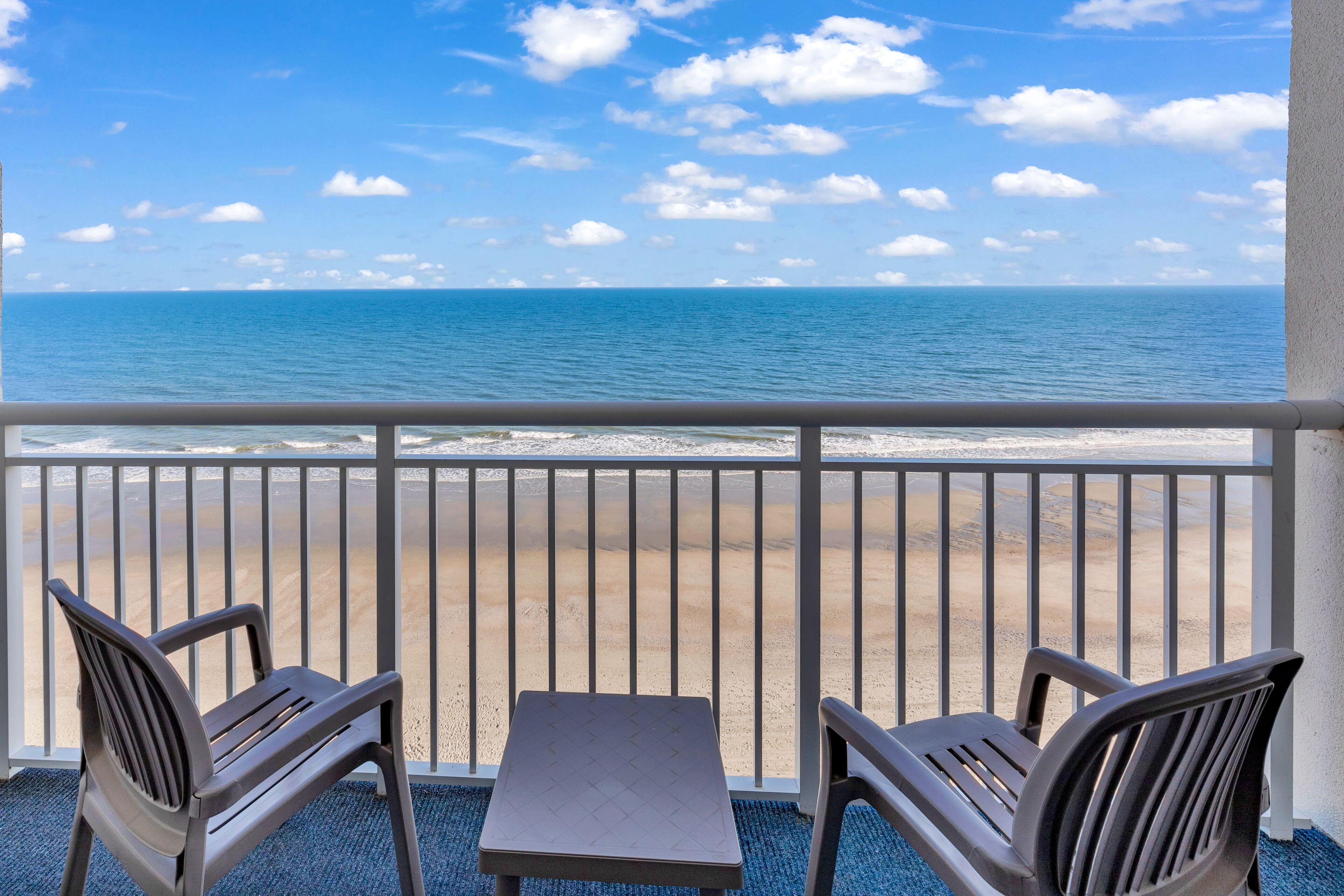 A balcony with two chairs and a table, overlooking a beach with a clear sky.