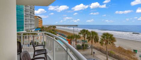 A balcony with a view of the beach, a pier, and a parking area with cars.