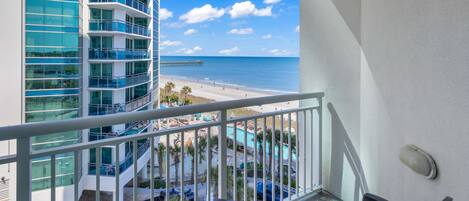 A balcony with a view of a beach and a pier, featuring a pool area and a multi-story building with balconies.