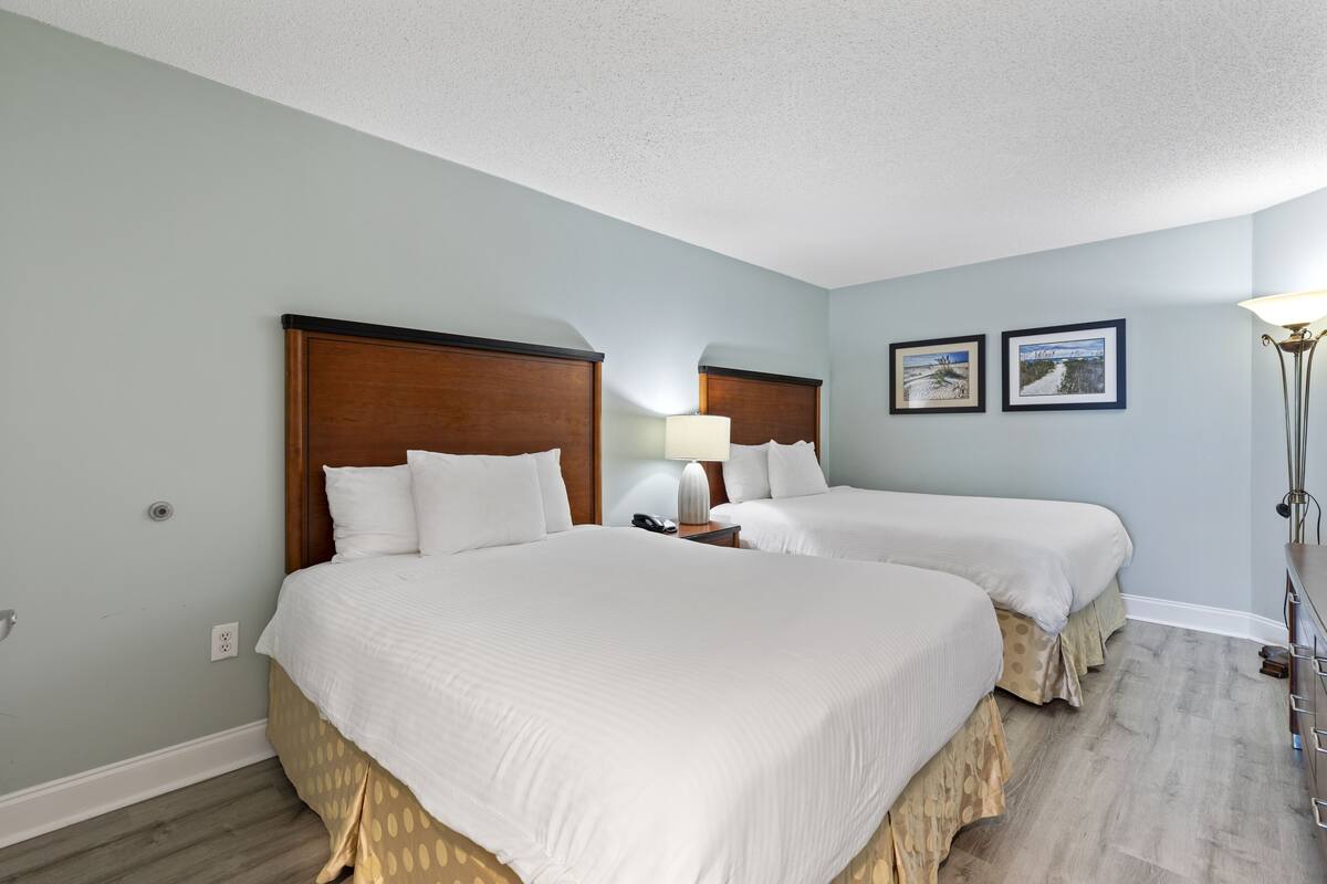 Habitación de hotel con dos camas, una mesita de noche con lámpara y dos cuadros en la pared.