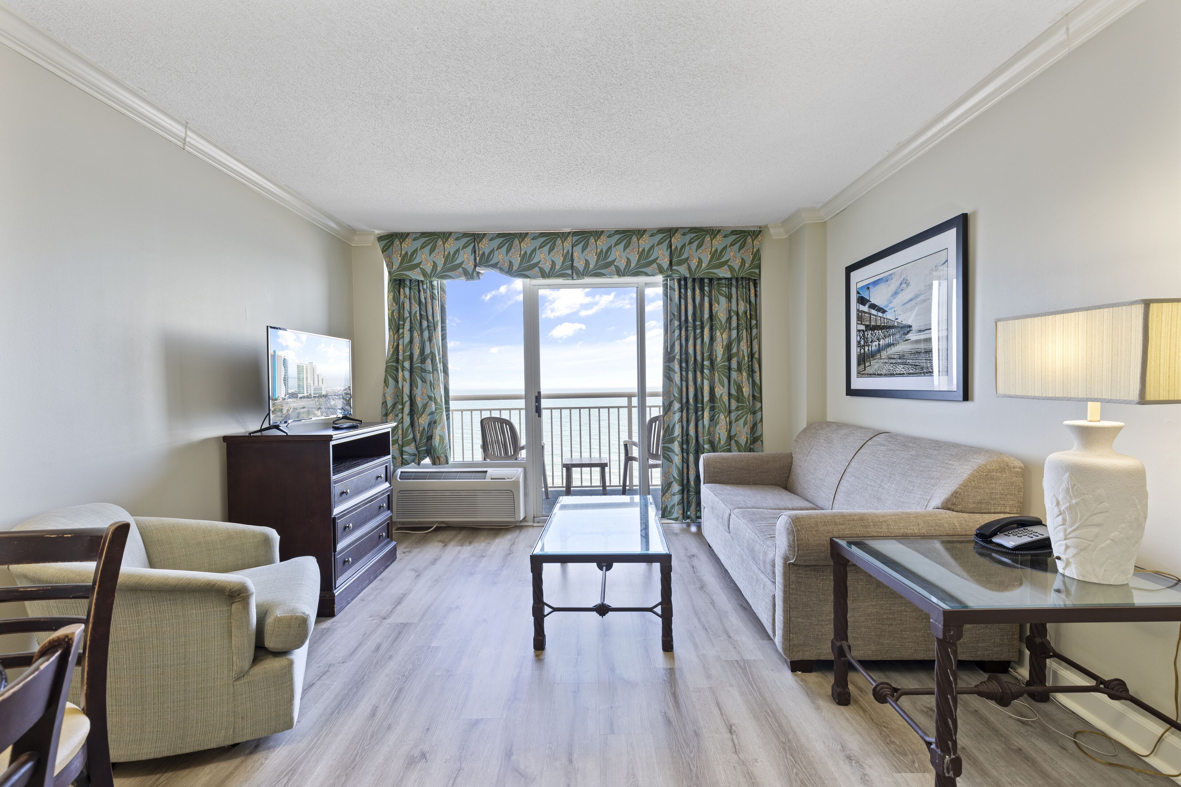 A hotel room with a balcony, a sofa, a chair, a coffee table, and a TV.