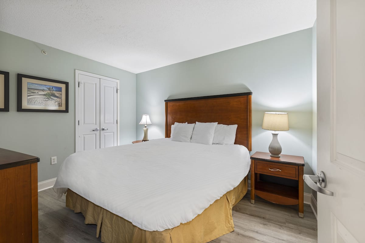 Un dormitorio con una cama grande, una mesita de noche con lámpara, un aparador y una puerta.