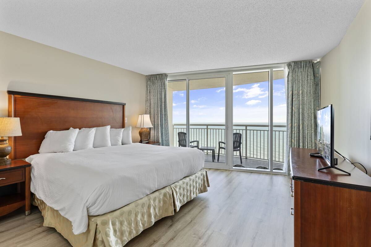Una habitación de hotel con una cama grande, un escritorio con televisor, un balcón con vista al océano y piso de madera.