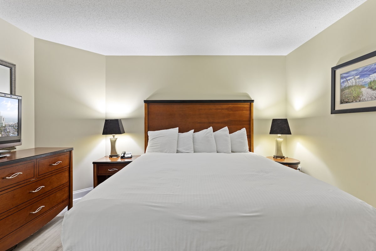 Habitación de hotel con una cama grande, cabecera de madera, dos mesitas de noche con lámparas, un tocador y un cuadro enmarcado en la pared.