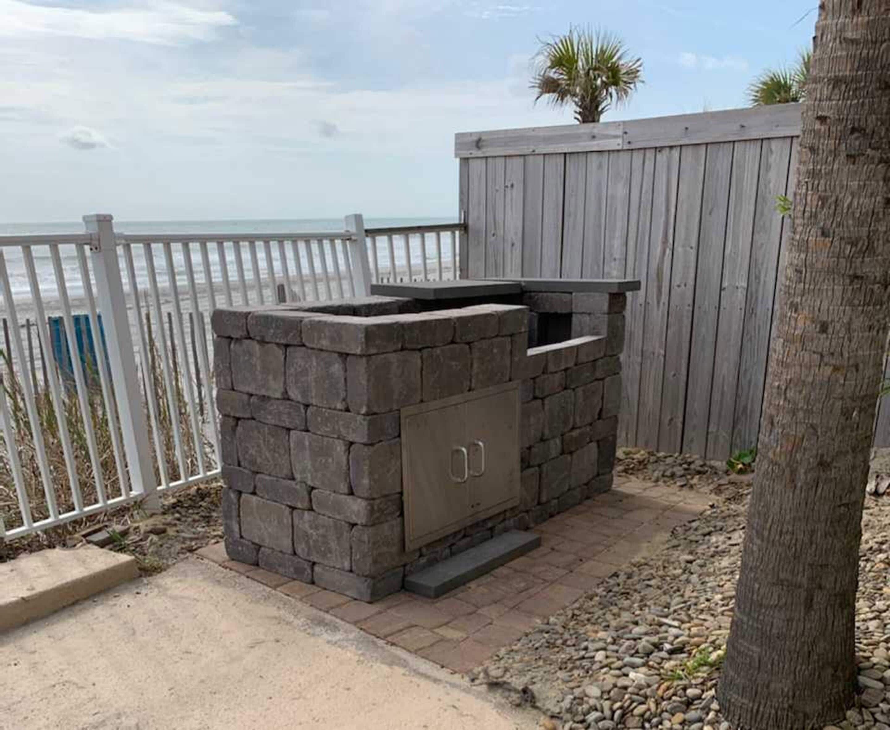 Una parrilla para barbacoa al aire libre con diseño de piedra, ubicada junto a una valla de madera y una palmera, con vista al océano de fondo.