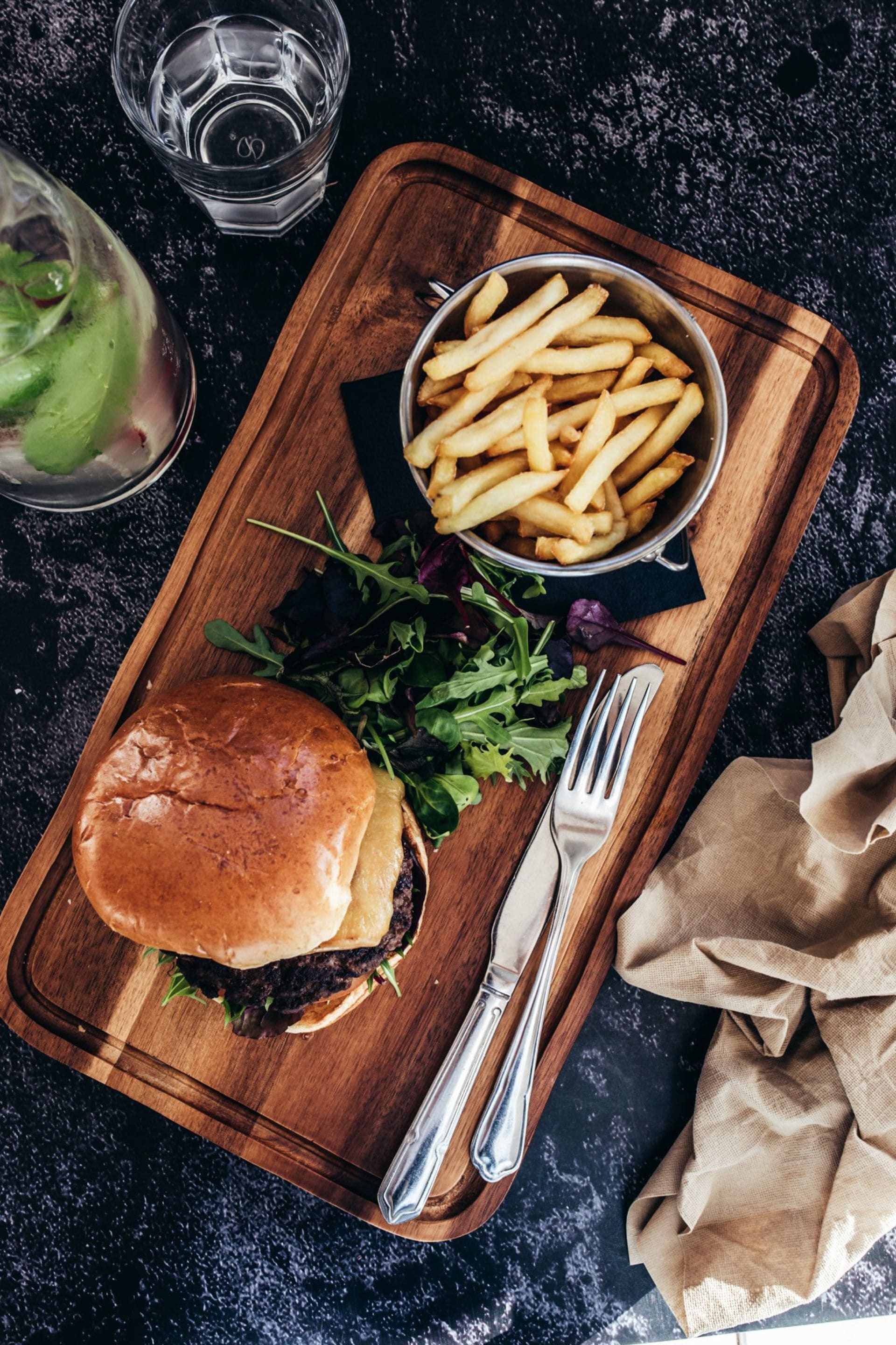 A wooden serving board with a burger, fries, and salad, accompanied by cutlery and a glass of water.