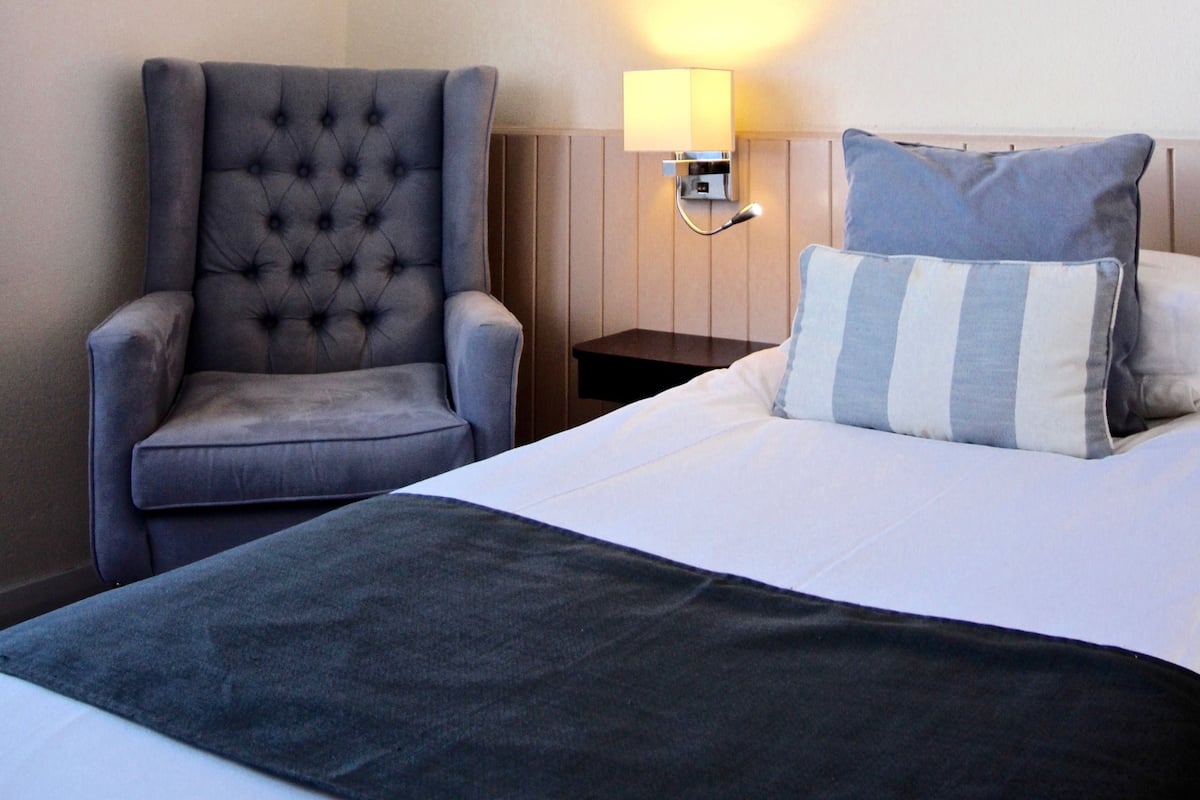 Habitación de hotel con una cama grande, un sillón capitonado, una mesita de noche con lámpara y un cojín a rayas.