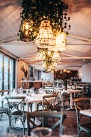 Interior de restaurante con mesas y sillas de madera, luces colgantes y vistas amplias a través de grandes ventanales.
