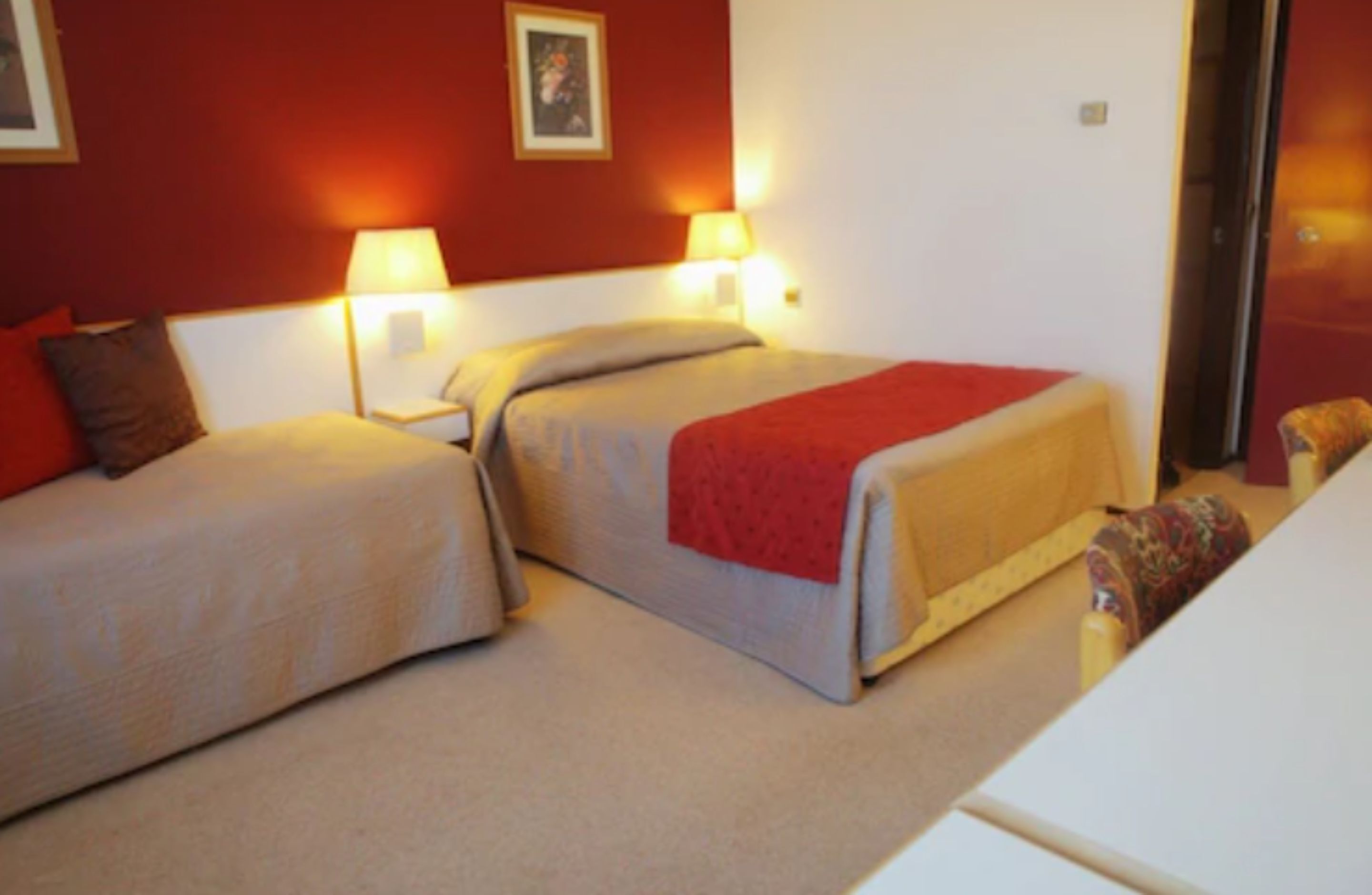 Quarto de hotel com duas camas, cada uma com um cobertor vermelho e luminárias laterais.