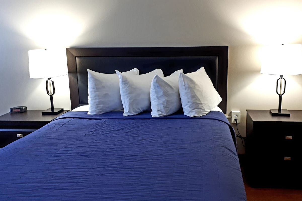 A neatly made bed with a dark blue bedspread and four white pillows. There are two bedside tables, each with a lamp and a small clock.