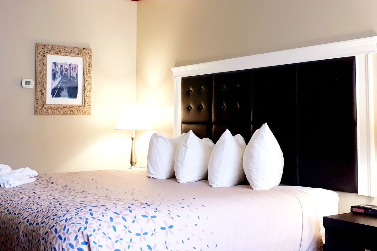 A hotel room with a bed, a bedside table, a lamp, a framed picture on the wall, and a thermostat.