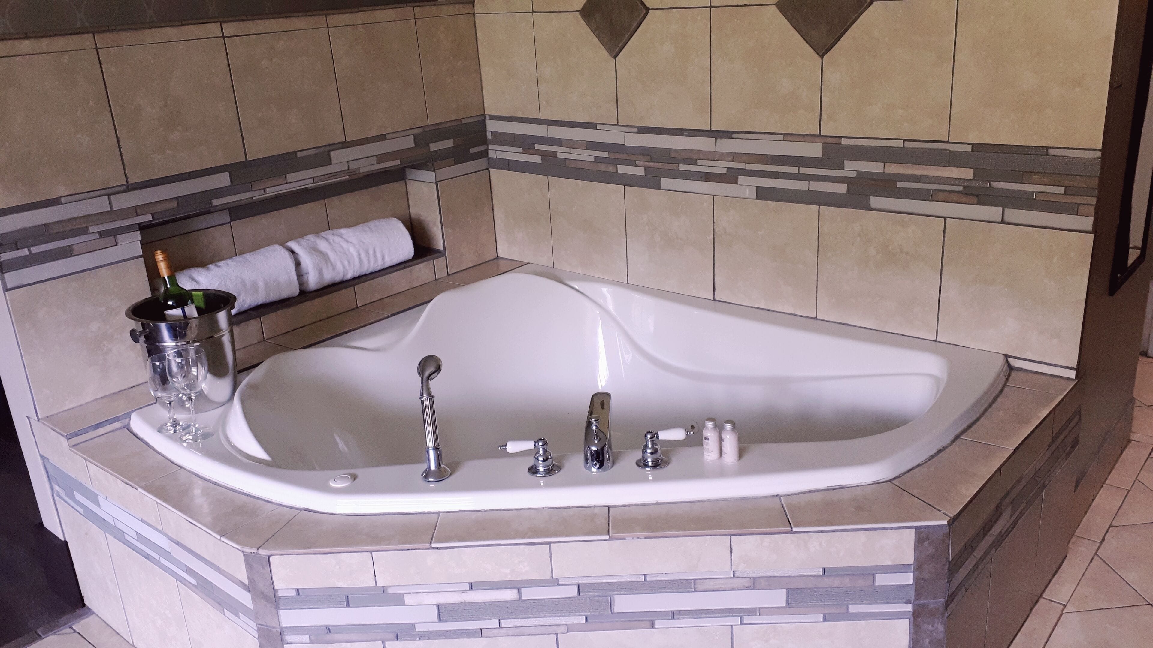 A large bathtub with a wine bottle and glasses on the side, a towel, and a bottle of champagne.