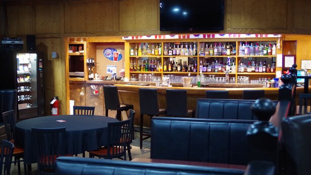 Una zona de bar con estantes de madera llenos de botellas de licores, un televisor montado en la pared y diferentes opciones de asientos, incluyendo mesas y cabinas.