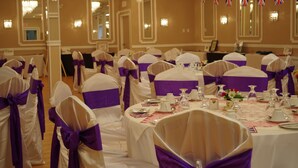 Un salón de banquetes con mesas redondas dispuestas para un evento formal, decorado con manteles blancos y cintas moradas.