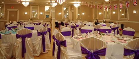 Un salón de banquetes con mesas dispuestas para un evento formal, decorado con cintas moradas y manteles blancos, y adornado con banderas británicas.