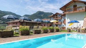 A poolside area with lounge chairs and umbrellas, a clear blue pool, and buildings with balconies in the background. - Hotel Alle Piramidi (Segonzano)
