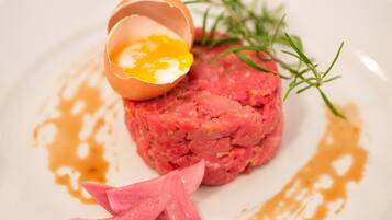 A dish with a raw meat patty, a cracked egg, and a sprig of rosemary on a white plate.