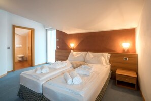 A hotel room with two beds, a wooden headboard, a bedside table, and a bathroom visible through an open door. - Hotel Alle Piramidi (Segonzano)