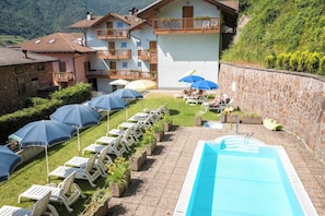 A poolside area with lounge chairs and umbrellas, a swimming pool, and a building with balconies. - Hotel Alle Piramidi (Segonzano)