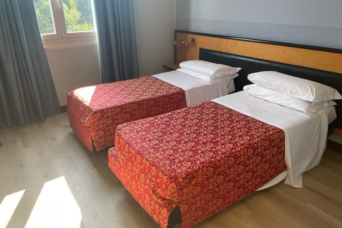 Zwei Einzelbetten mit roten, gemusterten Bettdecken, ein Holzkopfstück und ein Fenster mit Vorhängen.