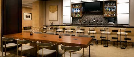 Una sala de conferències amb una taula de fusta llarga i diverses cadires, una zona de bar amb prestatgeries plenes de botelles, i una televisió penjada a la paret.