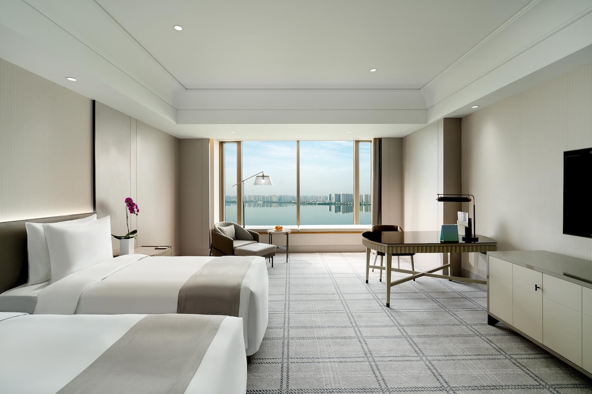 Habitación de hotel moderna con una cama grande, un escritorio, una silla y vistas a un paisaje urbano a través de amplios ventanales.