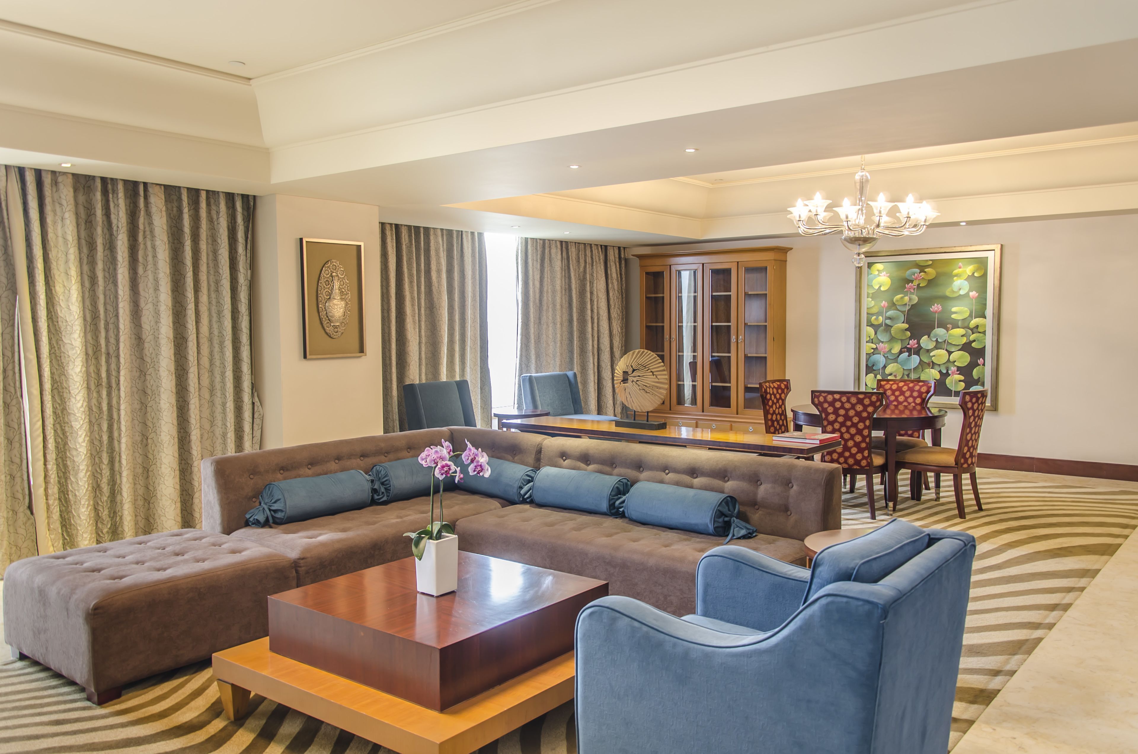 A modern hotel room with a sectional sofa, a wooden coffee table, and a dining area with chairs and a table.
