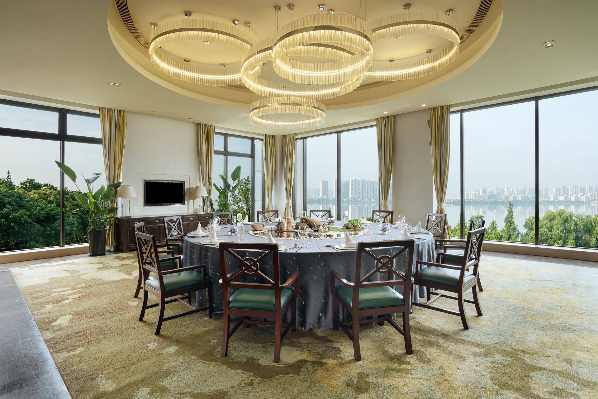 A dining room with a round table set for six, surrounded by chairs, a television on the wall, and a city view through large windows.