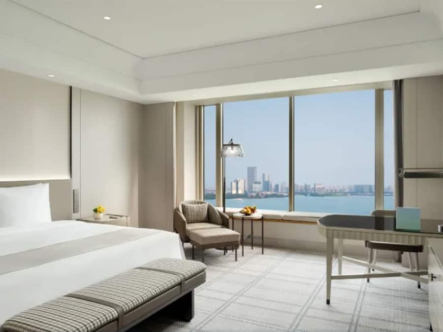 A modern hotel room with a large bed, a desk, a chair, and a view of the city skyline.