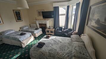 Habitación de hotel con dos camas individuales, un sofá, un televisor, una mesita de noche con lámpara y una silla.