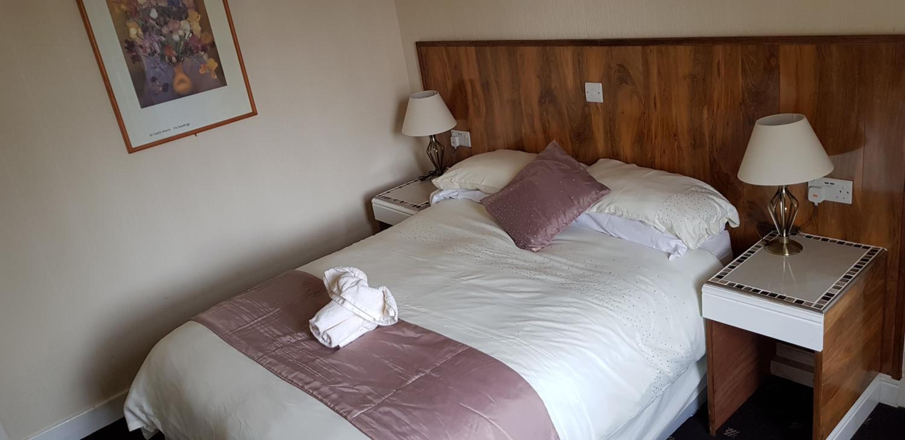 Een net opgemaakt bed met een roze kussen, twee bedlampjes en een ingelijst beeld aan de muur.